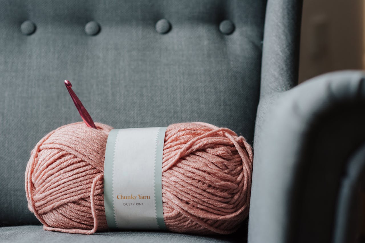 A ball of chunky dusky pink yarn with a crochet hook resting on a grey chair, perfect for crafts.